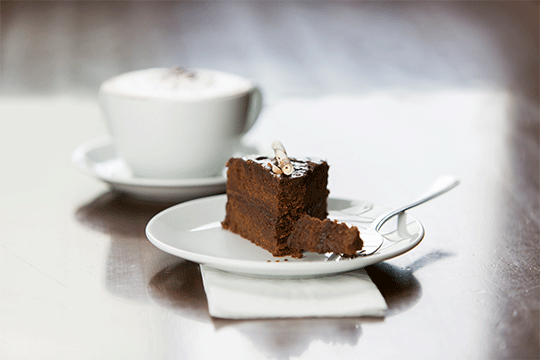 A slice of chocolate cake on a plate with a fork with a cup of cappuccino with chocolate on top.