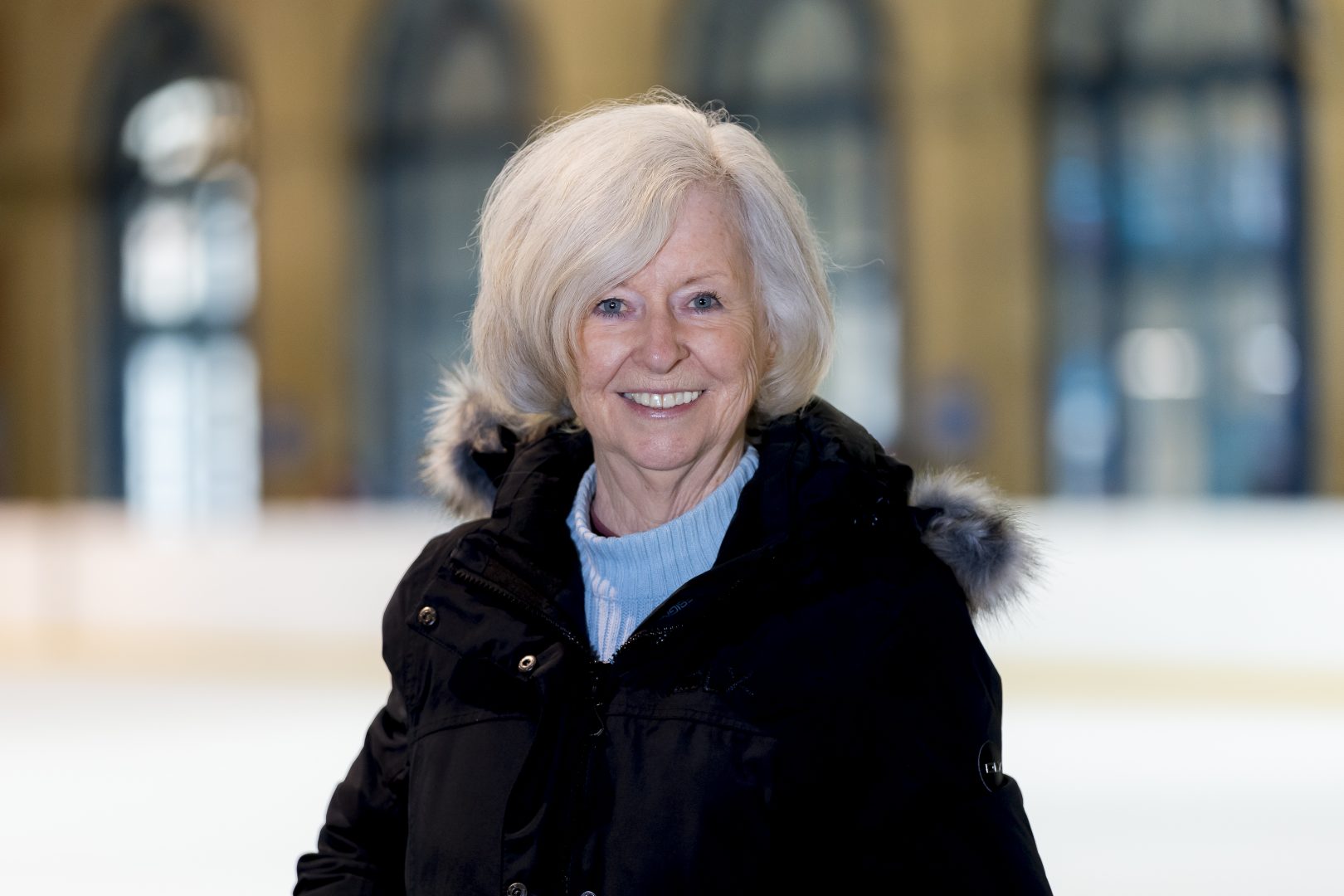 Woman with light hair, blue jumper and dark coat smiling whilst stood on an ice rink