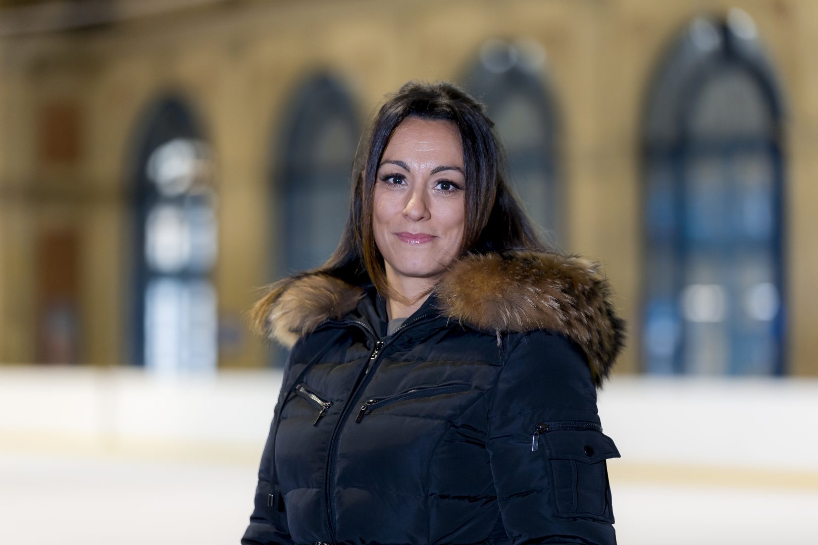 Woman with brown hair and black coat with fur collar smiles at camera whilst standing on an ice rink