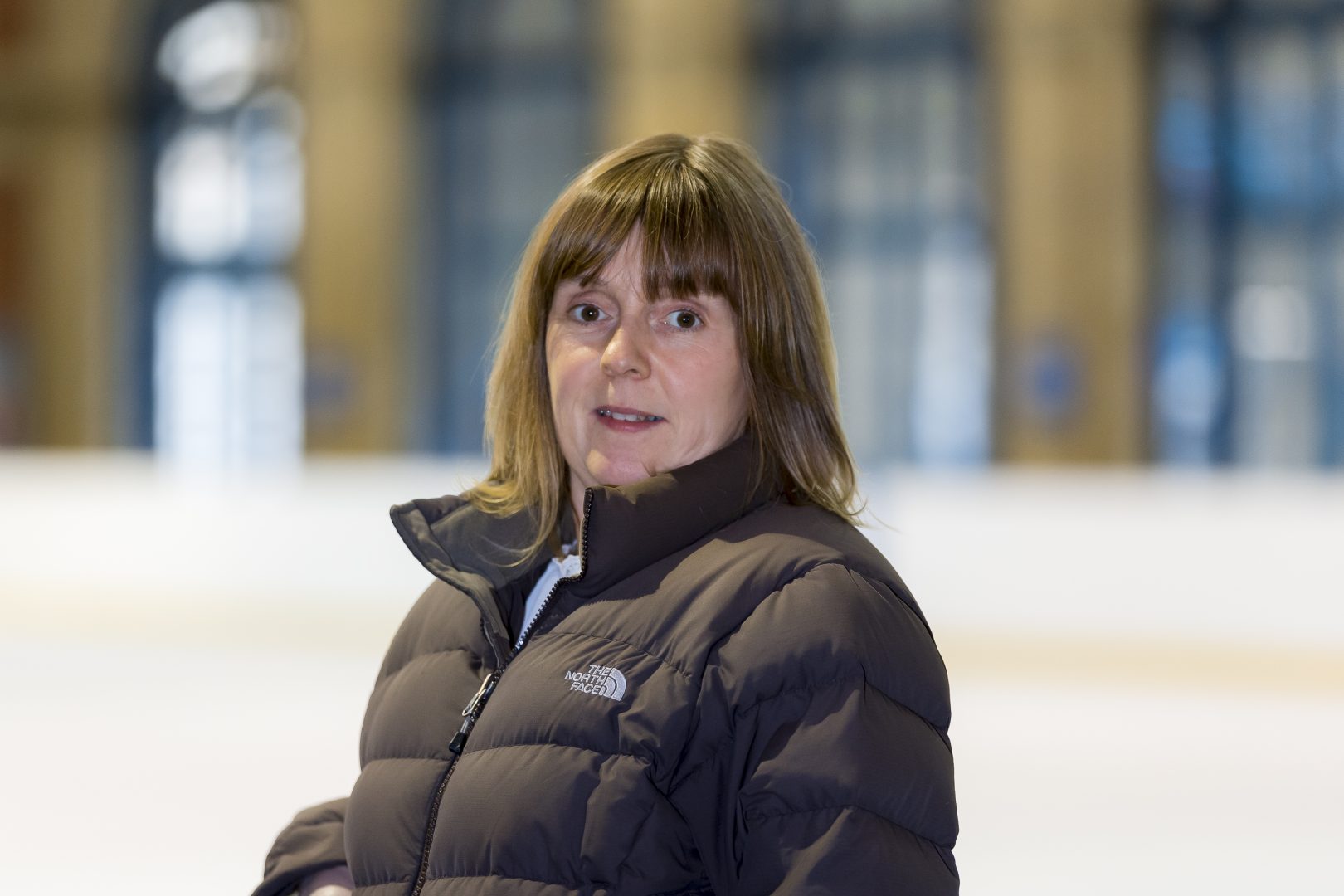 Woman with brown hair and black puffer jacket stands on an ice rink smiling