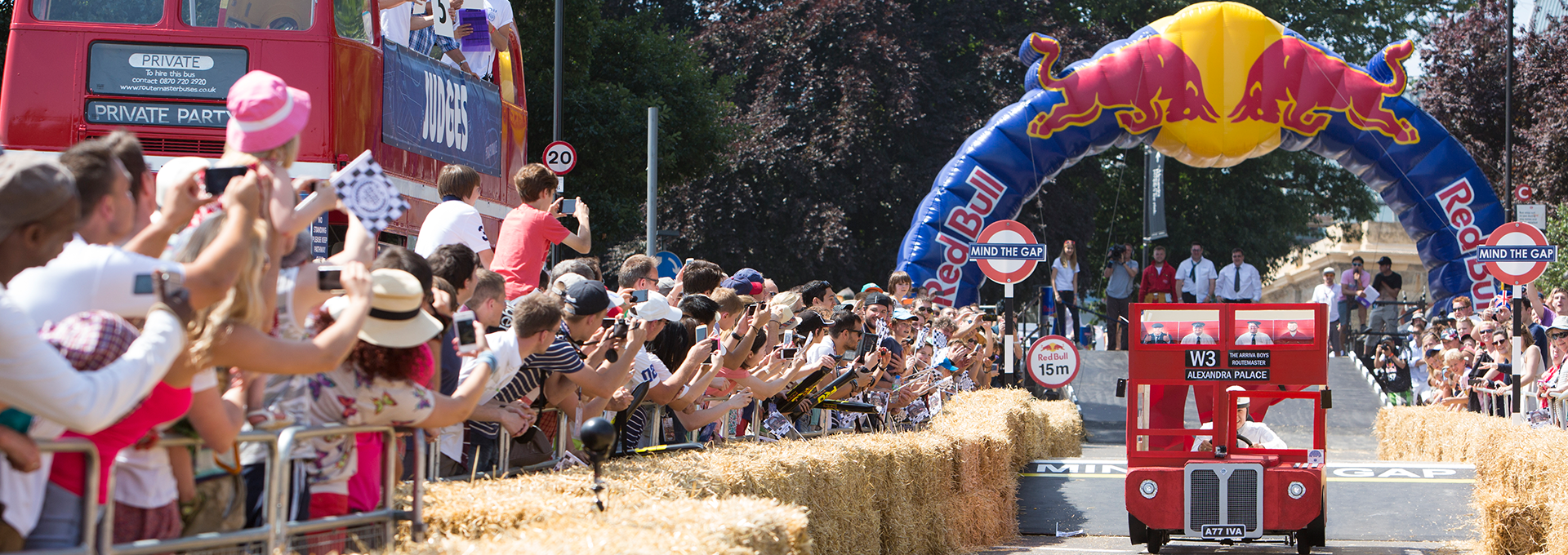 Play red bus being driven under a Red Bull archway finish line whilst people look on cheering and clapping.